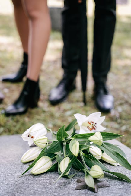 A bouquet of white lilies with open blossoms and closed buds, featuring long, green leaves, lies on a stone surface in the foreground. In the background, a person wearing black shoes and dark trousers