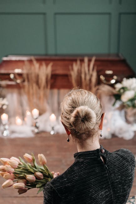A woman with her blonde hair styled in a neat bun, dressed in a black textured top, is holding a bouquet of pink tulips with long green stems. She is facing a wood-paneled wall decorated with candles 