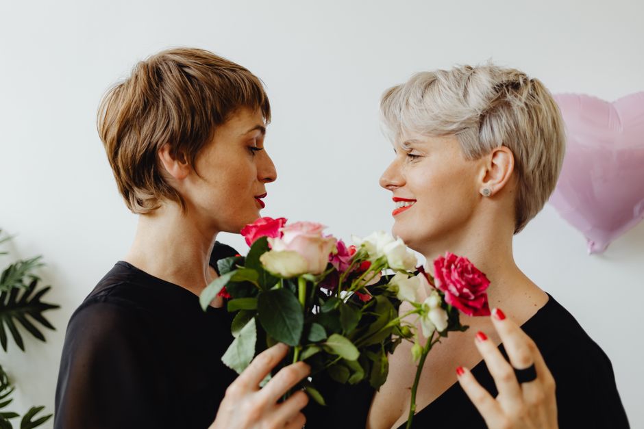 Two women with short, styled hair and red lipstick share a tender moment as one presents a bouquet of fresh flowers to the other. The bouquet features pink and white roses with green foliage, wrapped