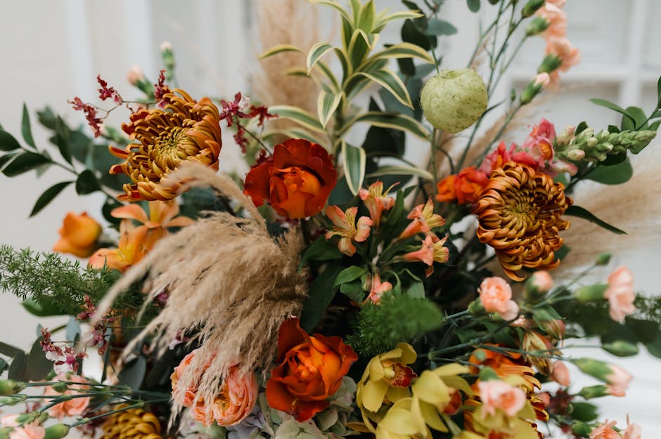 A vibrant floral arrangement featuring orange roses, large rust-colored chrysanthemums, and pink snapdragons interspersed with green foliage, including variegated leaves and sprigs of fern. The bouque