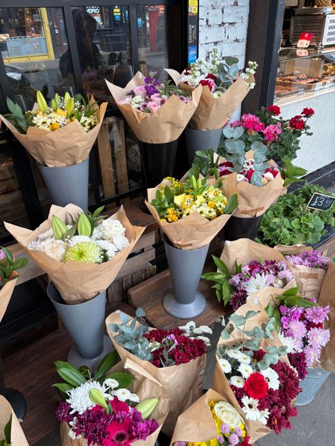 A display of multiple floral arrangements in tall, elegant grey vases and wrapped bouquets, featuring a variety of fresh flowers in vibrant colours. The arrangements include white lilies, yellow and p