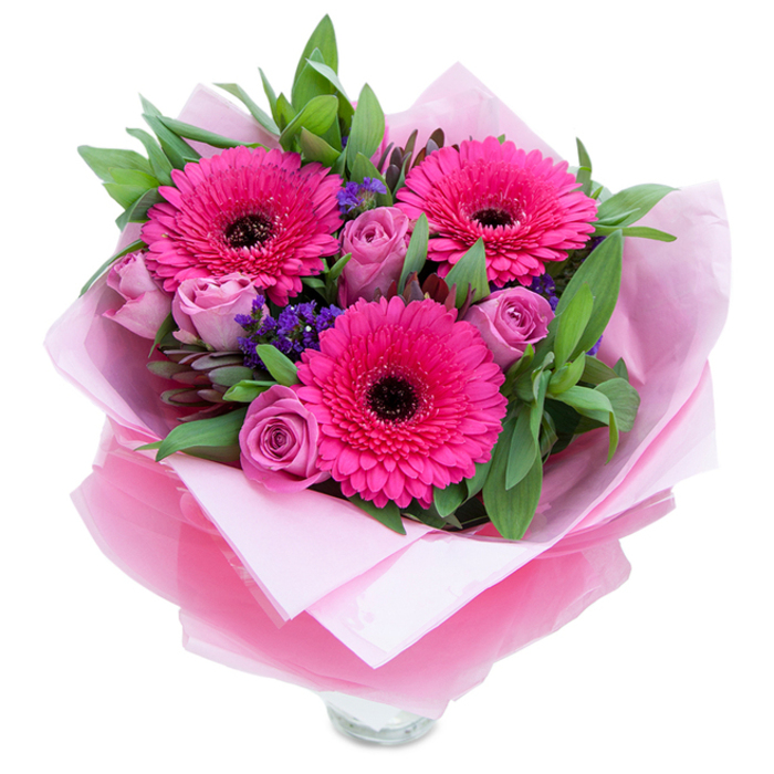 The charm of this bouquet is the way contrasting textures and a single, confident colour story come together: three fuchsia Gerbera daisies open like small suns, their radiating petals surrounding deep, almost black centres that anchor the eye. Interspersed among these are roses in a muted lavender-pink - each one unfurling with layered, velvety curls that soften the composition and lend a romantic hush. Little clusters of purple statice tuck into the spaces between blooms, their papery, slightly crisp texture introducing a rustic, wildflower note against the more polished petals.

Greenery forms a generous backdrop: broad glossy leaves that resemble salal or eucalyptus create fullness and offer a cool, leafy scent that balances the sweetness of the roses. The blooms are enveloped in a pale pink tissue paper inner wrap, which feels soft and matte to the touch, while an overlay of translucent fuchsia-pink cellophane with a slight crinkle gives the bouquet a subtle, decorative shimmer. The top rim of a clear glass vase is visible beneath, a reassuring detail that indicates the flowers are arranged for immediate display and fresh water.

I imagine presenting this arrangement from a local Camberwell florist's table - perhaps delivered along Camberwell Church Street or Bellenden Road - where the colours brighten a mantelpiece or bring a warm, celebratory tone to a small gathering. It's suited to birthdays, anniversaries, or a spontaneous gesture of affection, designed to feel intimate and thoughtfully made.