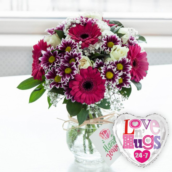A lush, celebratory bouquet arranged by local hands in Camberwell fills a clear glass vase, the scene framed by bright, diffused light that suggests a window looking out towards Camberwell Green or one of the nearby residential streets. Dominating the composition, several impressive gerbera daisies in a vivid magenta-fuchsia hue open wide, each with a deep, almost espresso-coloured centre that anchors the colour palette. Around these statement blooms, clusters of two-toned chrysanthemums create a captivating pattern, their petals dark purple near the centre, fading smoothly to clean white tips, circling around bright yellow disks that feel almost like little lanterns. Among the bolder tones, creamy white roses are tucked in gracefully, their petals satin-smooth and only just beginning to swirl open, offering a touch of timeless elegance. Airy sprays of white gypsophila drift in and out of the composition like fine lace, softening edges and filling any gaps so the bouquet appears generously full and rounded. Fresh green foliage peeks from beneath and between the flowers, its varied textures - from broad, glossy leaves to lighter, feathery fronds - adding depth and a natural, freshly picked feel. The entire arrangement is gathered into a stout, rounded glass vase filled with clear water, the carefully trimmed stems clearly visible, reflecting the skilled craftsmanship expected from a trusted neighbourhood florist like Camberwell Flowers. A length of rustic natural twine is tied neatly around the vase neck, supporting a small rectangular white tag printed with a simple pink rose motif and friendly green script, echoing a hand-written dedication. To the right side, a shiny foil heart-shaped balloon with a reflective silver edge and white centre sits slightly forward, emblazoned with the phrase Love n' Hugs 24-7 in playful, patterned lettering across shades of pink, purple and blue. The softly blurred, light-filled background enhances the joyful mood, making this image an ideal representation of a romantic surprise, birthday treat, or just-because bouquet ready for local delivery across Camberwell and the surrounding SE5 streets.