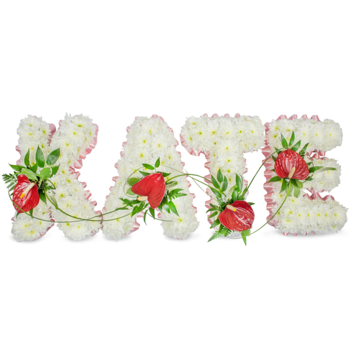 A beautifully crafted memorial arrangement spells out KATE in large, raised letters composed entirely of small, pristine white spray chrysanthemums, their dense heads forming a smooth, cloud-like surface that suggests peace and reverence. The outer edge of each letter is softly defined by a light-pink pleated ribbon that gives a faint blush contrast to the white and provides a polished, respectful finish. Scattered within the chrysanthemum field are vivid red anthurium blooms - glossy, heart-shaped spathes with noticeable spadices - positioned to act as warm, loving accents against the serene white. Interspersed greenery, from broad glossy leaves to delicate ferny fronds and fine trailing vines, is arranged to weave through the letters and to link the composition visually, adding depth, texture and a living, natural rhythm. The arrangement sits on a clean white background with a slight drop shadow beneath each letter, lending a sculptural, three-dimensional quality that reads clearly from a distance in a chapel or viewing room. The tactile mix - the soft, almost powdery chrysanthemum surface alongside the sleek, waxy anthuriums and crisp foliage - creates a quiet sensory memory, as if one could smell the cool green notes and feel the layered textures. Intended as a sympathetic, bespoke tribute for funeral services and commemorations in Camberwell, near Rye Lane and the churches around Denmark Hill, this piece conveys careful workmanship, dignity and heartfelt remembrance.